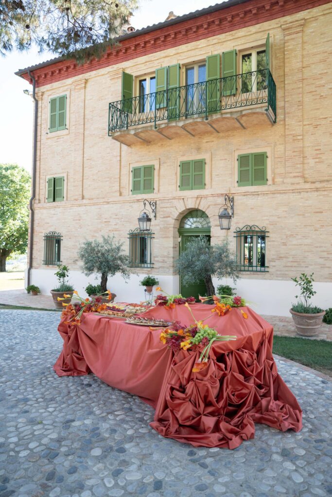 Hochzeit im Maniero Montecassiano: Euer Hochzeitswochenende in einer Boutique-Villa in den Marken 49 Ein Buffettisch mit einem langen, drapierten orangefarbenen Tischtuch im Freien auf dem Maniero Montecassiano.