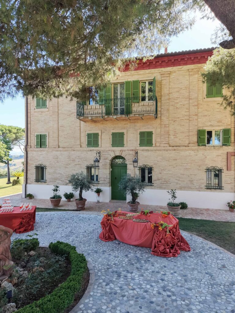 Hochzeit im Maniero Montecassiano: Euer Hochzeitswochenende in einer Boutique-Villa in den Marken 11 Eine Steinvilla in Maniero Montecassiano mit grünen Fensterläden und Tischen auf einem gepflasterten Innenhof.