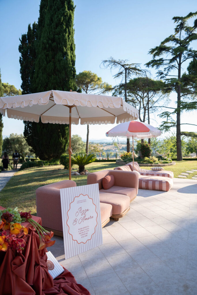 Hochzeit im Maniero Montecassiano: Euer Hochzeitswochenende in einer Boutique-Villa in den Marken 7 Außenbereich im Maniero Montecassiano mit einem Pizza & Amore-Schild, Sitzgelegenheiten und gestreiften Sonnenschirmen.
