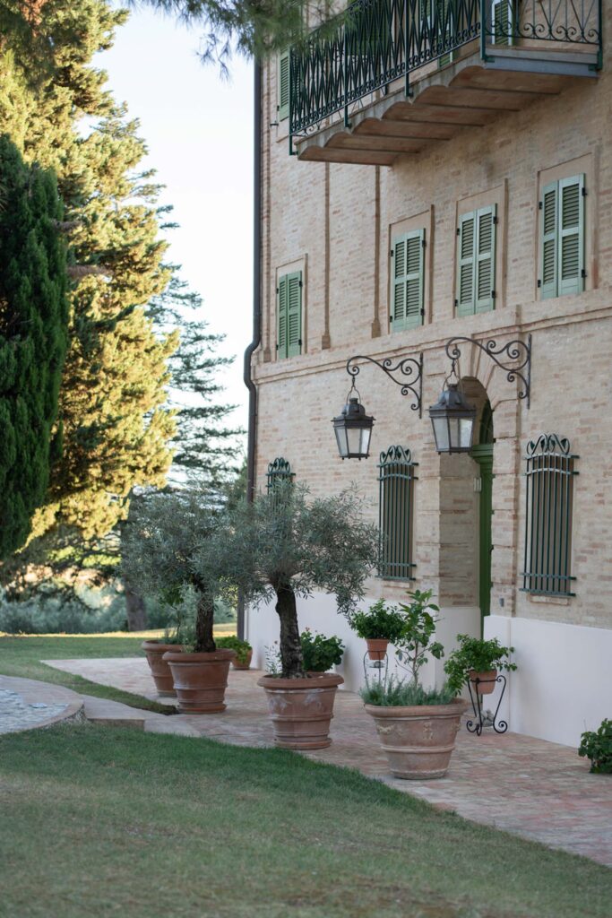 Hochzeit im Maniero Montecassiano: Euer Hochzeitswochenende in einer Boutique-Villa in den Marken 54 Ein Backsteinhaus im Stil von Montecassino mit grünen Fensterläden und Olivenbäumen in Töpfen auf einer Terrasse.