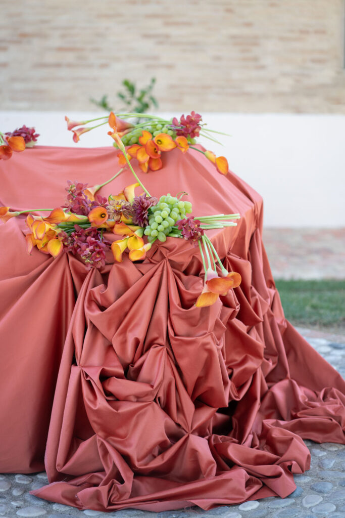 Hochzeit im Maniero Montecassiano: Euer Hochzeitswochenende in einer Boutique-Villa in den Marken 50 Eine korallenfarbene Tischdecke von Maniero Montecassiano, auf der orangefarbene Calla-Lilien und grüne Weintrauben drapiert sind.