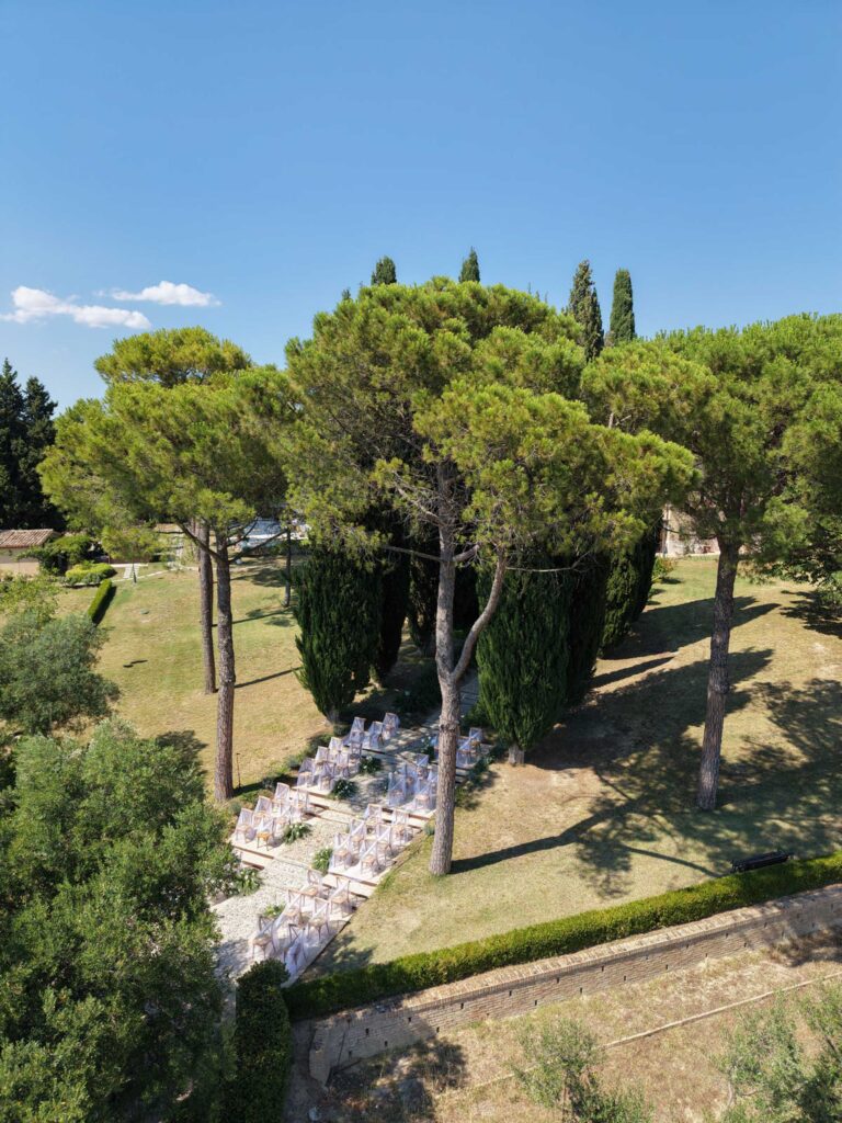 Hochzeit im Maniero Montecassiano: Euer Hochzeitswochenende in einer Boutique-Villa in den Marken 28 Reihen weißer Hochzeitsstühle, aufgestellt für eine Hochzeit in Marken in einem Garten zwischen hohen Kiefern.