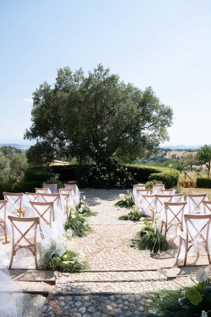 Hochzeit im Maniero Montecassiano: Euer Hochzeitswochenende in einer Boutique-Villa in den Marken 41 Stühle, die für eine Hochzeitszeremonie in einer Boutique-Villa in Italien entlang eines Weges mit Blick auf einen großen Baum aufgestellt wurden.