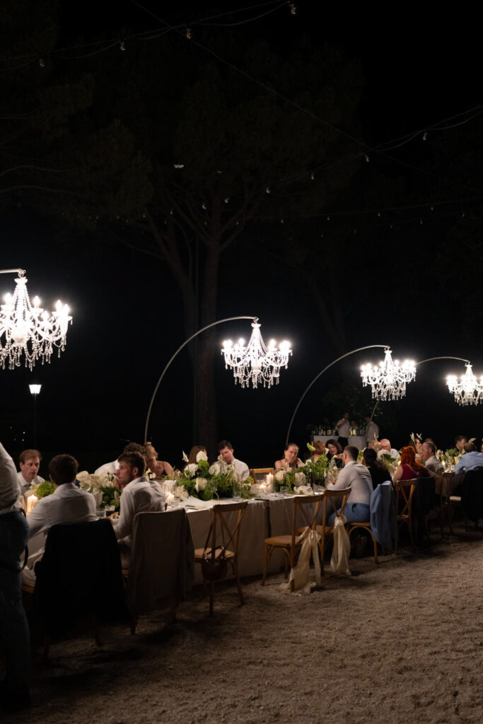 Hochzeit im Maniero Montecassiano: Euer Hochzeitswochenende in einer Boutique-Villa in den Marken 45 Gäste speisen an einem langen Tisch im Freien, der von Kristallkronleuchtern beleuchtet wird, bei der Hochzeit im Maniero.