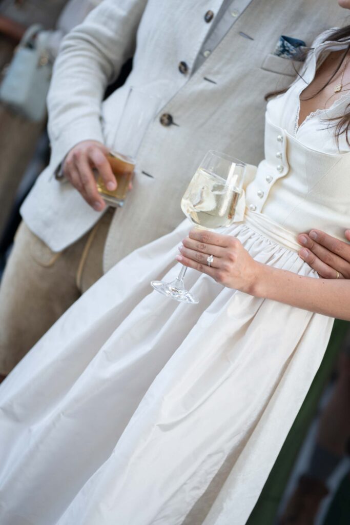 Eine Braut in einem weißen Kleid, die ein Glas Champagner in der Hand hält und ihren Arm um den Bräutigam gelegt hat, im Rosewood Schloss Fuschl.