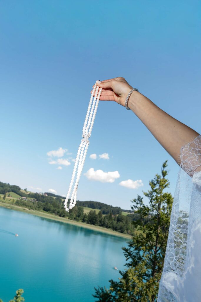 Bei einer Hochzeit auf Schloss Fuschl mit Blick auf den See hält eine Hand eine dreisträngige Perlenkette in der Hand.