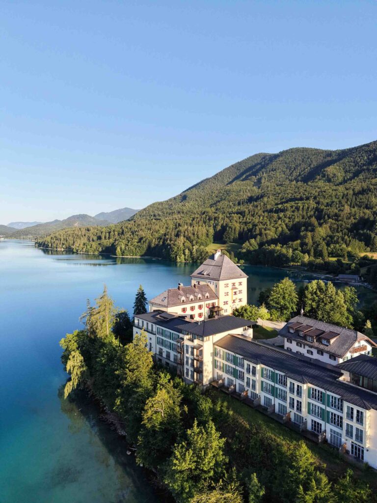 Das Rosewood Schloss Fuschl am Ufer eines blauen Sees, vor der Kulisse eines großen, bewaldeten Berges.