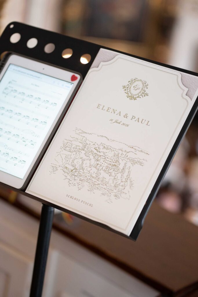 Das Hochzeitsprogramm für Elena und Paul auf Schloss Fuschl auf einem Notenständer neben einem Tablet.