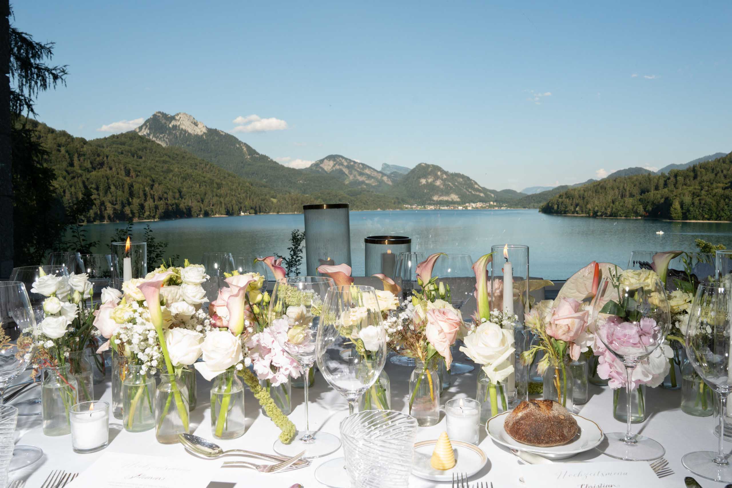 Ein gedeckter Tisch mit Blumenvasen und Gläsern mit Blick auf einen See und die Berge auf Schloss Fuschl.