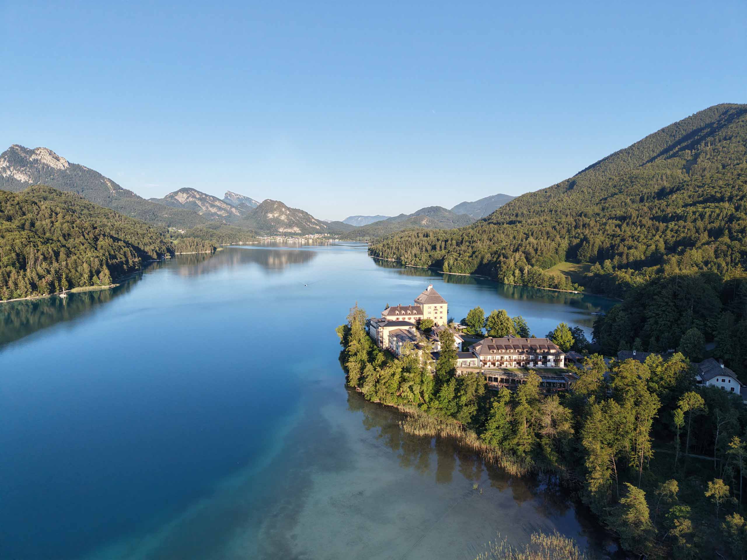 Das Rosewood Schloss Fuschl, ein Schlosskomplex auf einer bewaldeten Halbinsel, die in einen blauen Alpensee hineinragt.