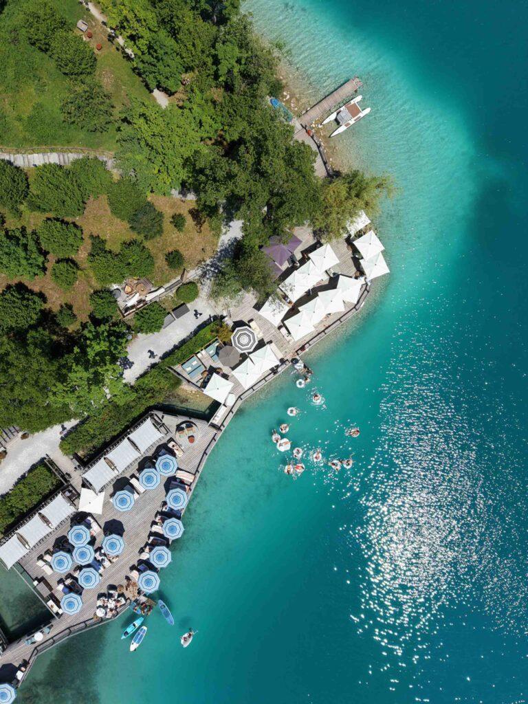 Luftaufnahme der Seeterrasse des Rosewood Schloss Fuschl mit Tischen, Sonnenschirmen und Badegästen.