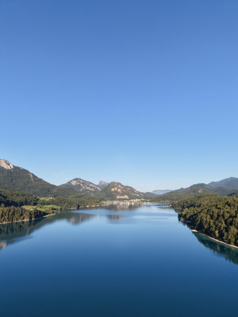Ein ruhiger See, umgeben von bewaldeten Bergen, am Rosewood Schloss Fuschl unter strahlend blauem Himmel.