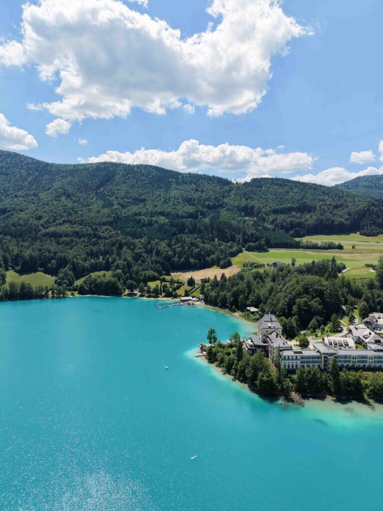 Luftaufnahme eines türkisfarbenen Sees beim Rosewood Schloss Fuschl, umgeben von Wald und Bergen.