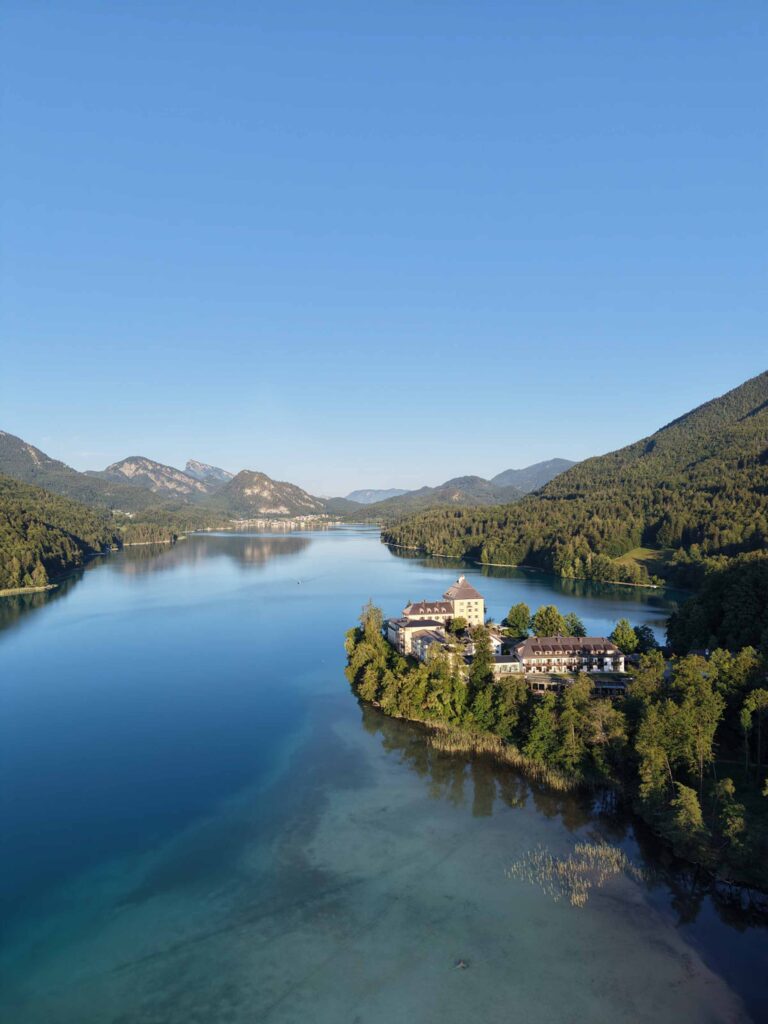 Das Rosewood Schloss Fuschl liegt auf einer bewaldeten Halbinsel in einem blauen See, umgeben von Bergen.