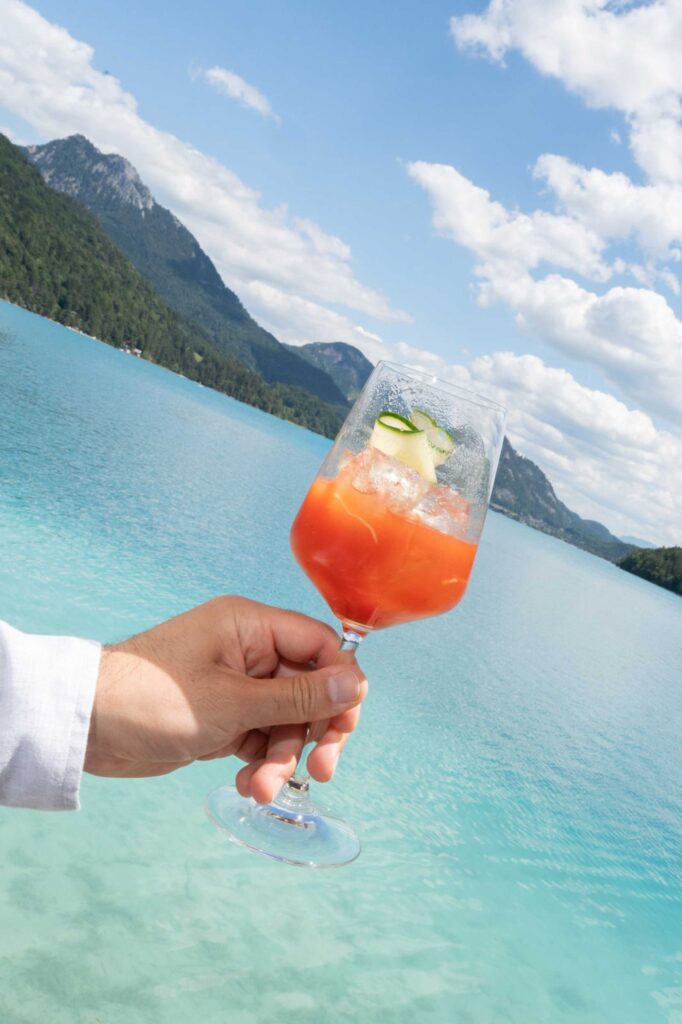 Im Rosewood Schloss Fuschl hält eine Hand einen roten Cocktail über einem türkisfarbenen See.
