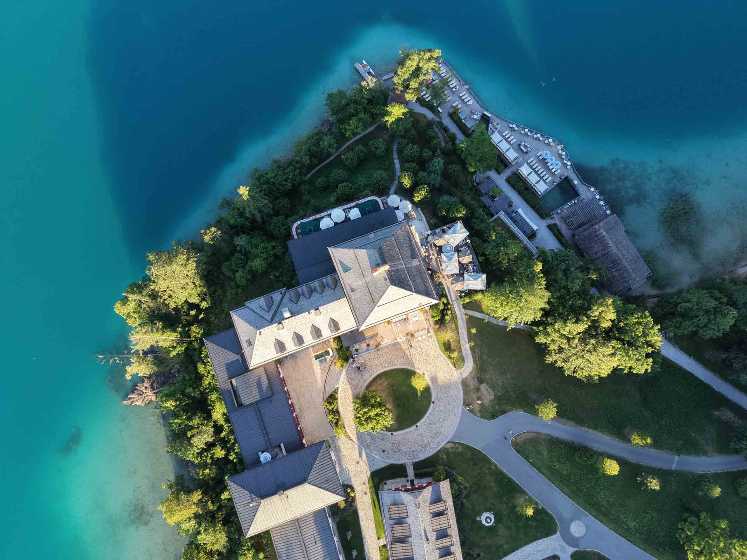 Luftaufnahme des Rosewood Schloss Fuschl auf einer bewaldeten Halbinsel, umgeben von türkisfarbenem Wasser.