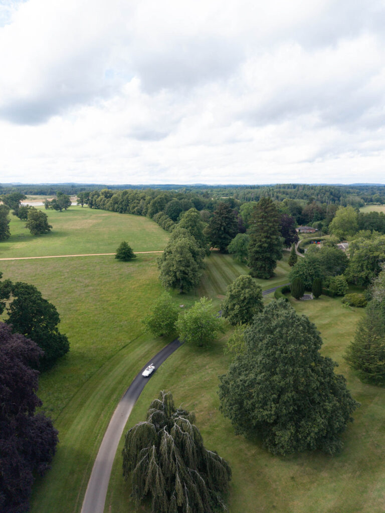 Ein weißes Cabrio, das eine kurvenreiche Zufahrt durch einen großen, grünen Park entlangfährt.